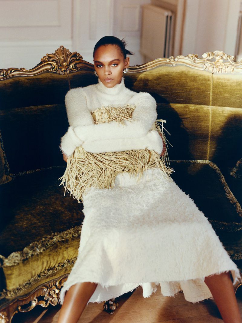 Elegant portrait of a woman with dark hair, posing on a velvet couch in a textured dress and sweater with unique straw embellishments.