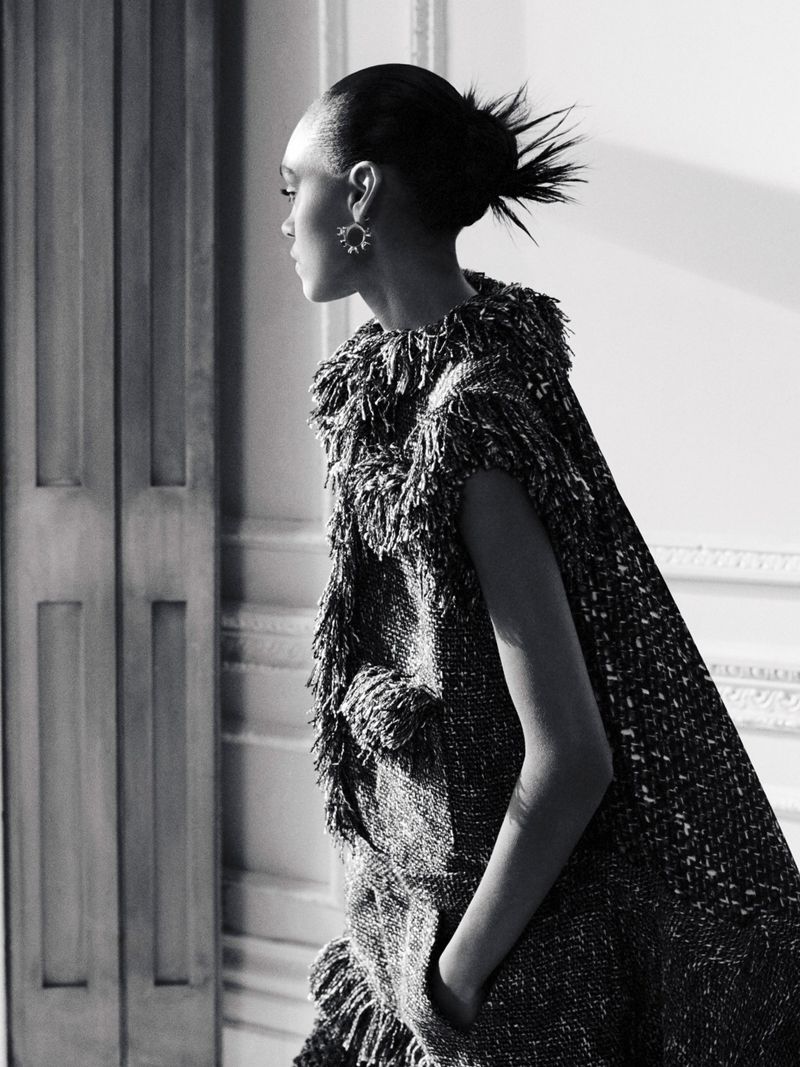 Black and white fashion shot of a woman in a fringed sleeveless dress looking away from the camera with a high-fashion hairstyle.