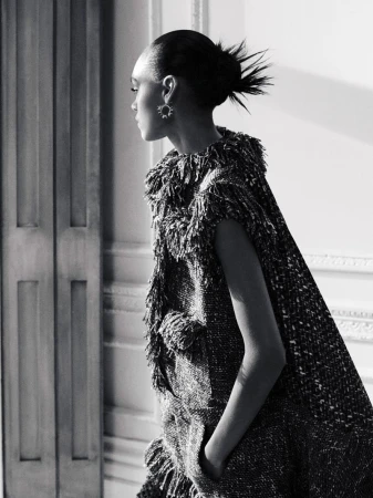 Black and white fashion shot of a woman in a fringed sleeveless dress looking away from the camera with a high-fashion hairstyle.
