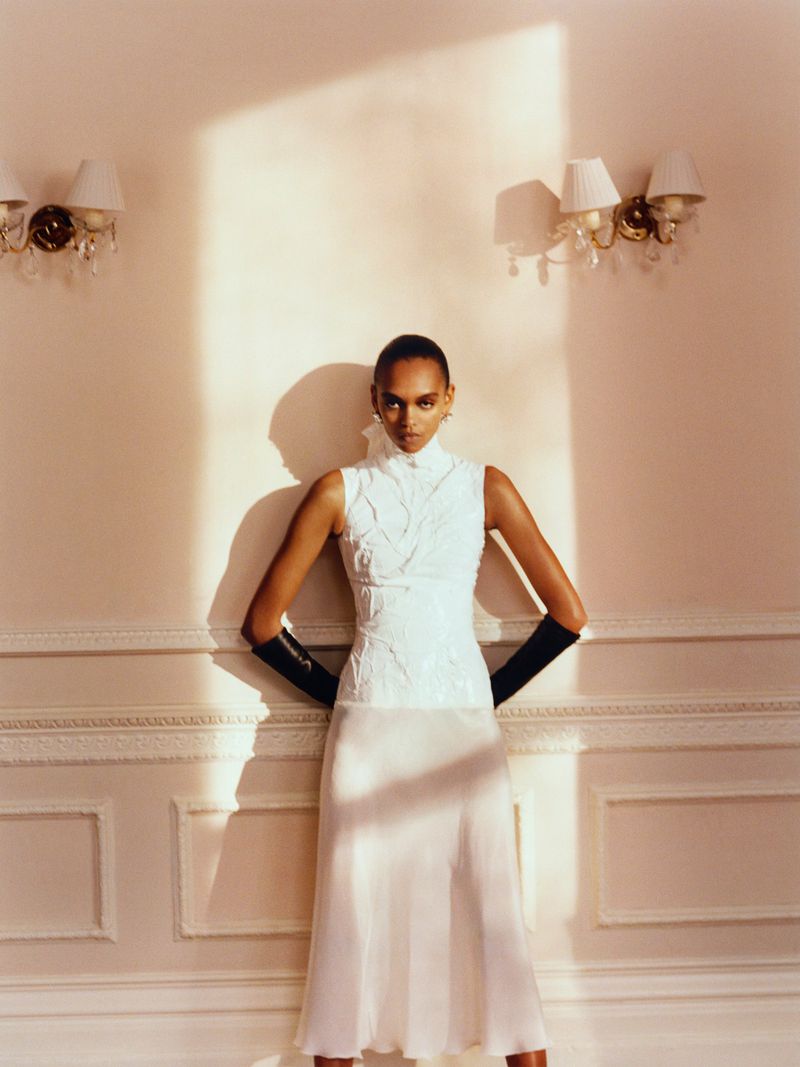 A dark-haired woman models a white top and skirt in a brightly lit interior with sconces on the walls and a high fashion, editorial feel.