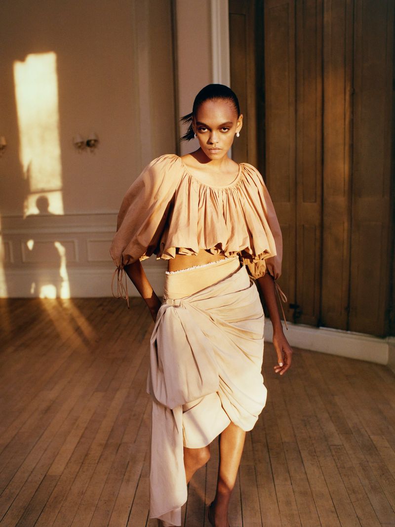 An adult woman with dark hair and bare feet poses in a tan outfit in a room with wood floors, illuminated by natural light from a window.
