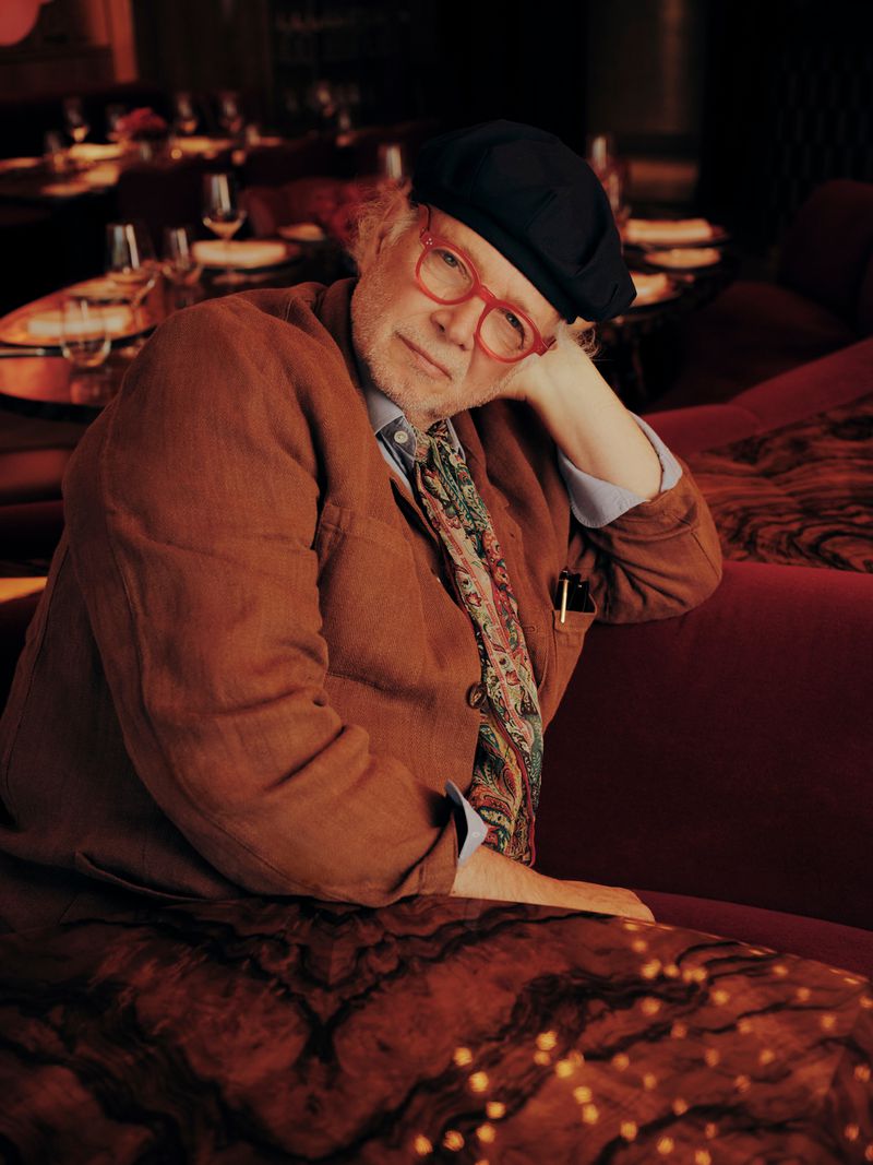 Stylish older man with glasses, brown blazer, and beret, sitting on a red patterned sofa in a restaurant, illuminated by warm lighting.