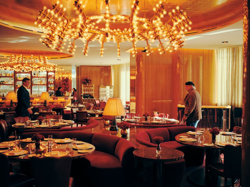 Elegant restaurant interior with tables set for dining, warm lighting from chandeliers, and people in the background at the bar.