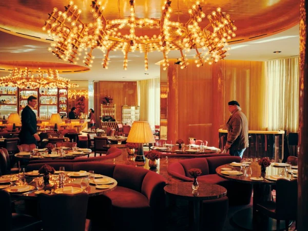 Elegant restaurant interior with tables set for dining, warm lighting from chandeliers, and people in the background at the bar.