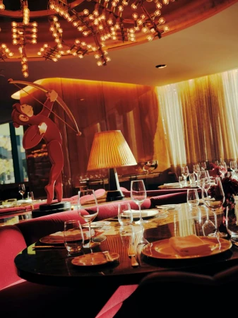 Elegant restaurant interior with a dining table set for service, a red Cupid statue, artistic light fixture, and warm golden lighting.