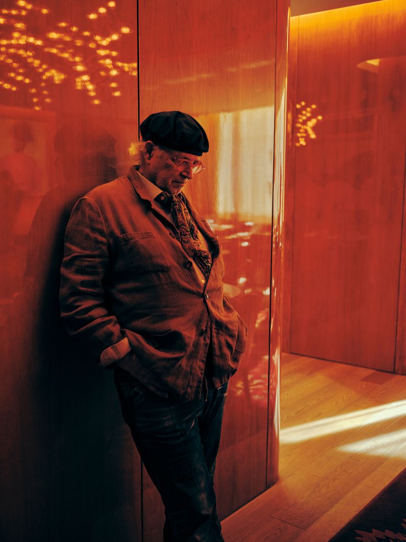 Elderly man in a beret leans against a wall in an indoor corridor with warm lighting, wearing a jacket, patterned scarf, and dark jeans.