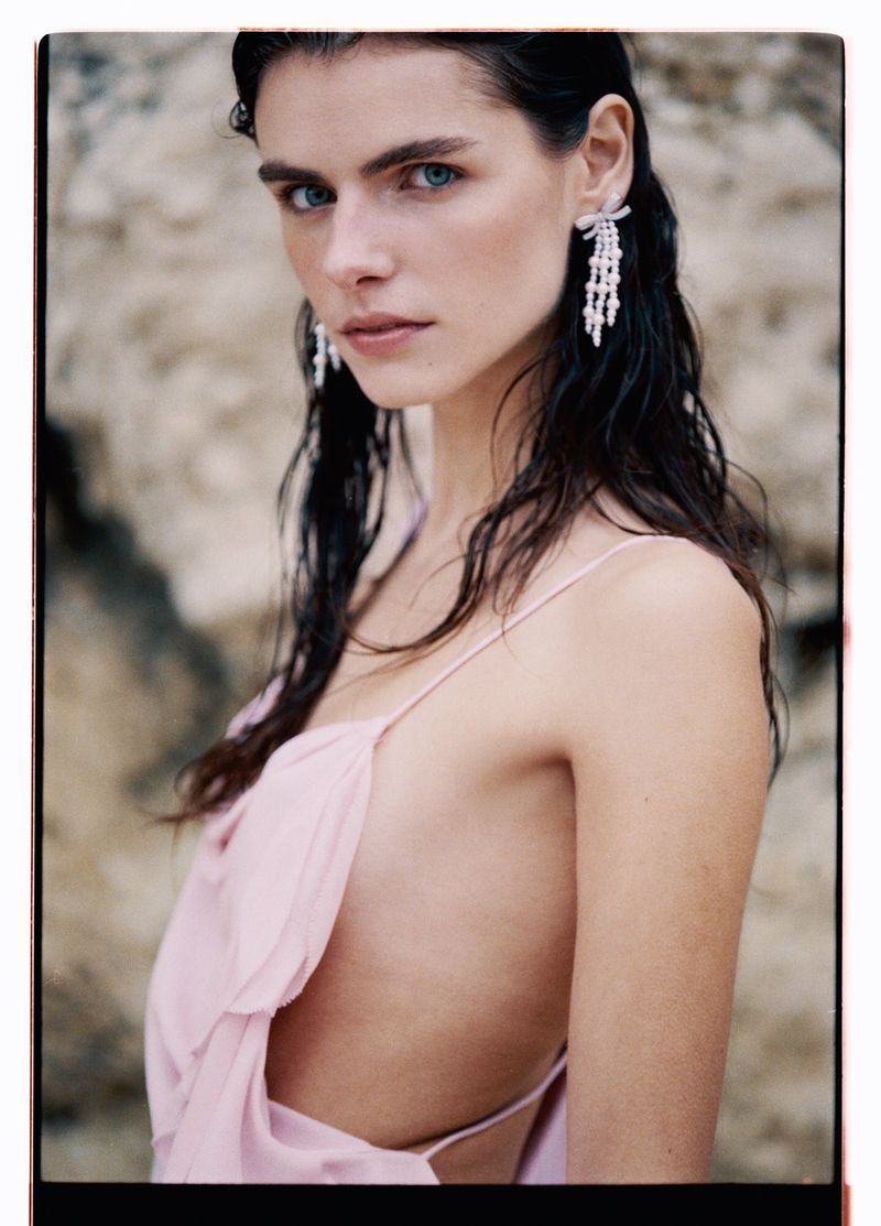 Portrait of a woman with wet dark hair wearing large crystal earrings and a pink gown, posing in front of a textured tan rock background.