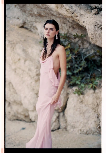 A woman in a pink dress poses in front of a large rock formation, wearing dangling earrings, showcasing fashion in a natural outdoor setting.