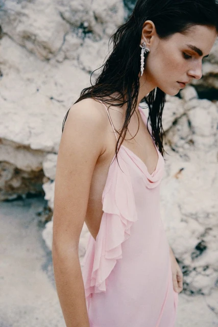 Elegant woman in a pink dress posing against a rocky background, wearing dangling earrings, looking down with wet black hair.