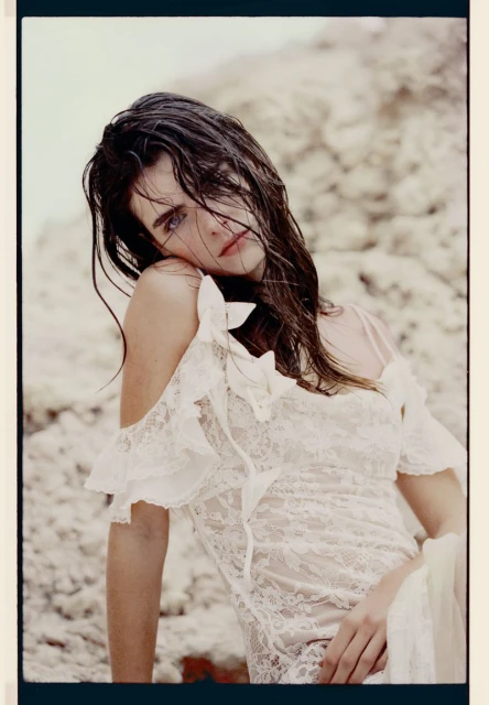 Brunette woman in a white lace dress gazes at the camera, posing near a light-colored rock formation in a fashion-style photo shoot.