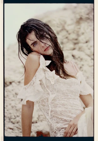 Brunette woman in a white lace dress gazes at the camera, posing near a light-colored rock formation in a fashion-style photo shoot.