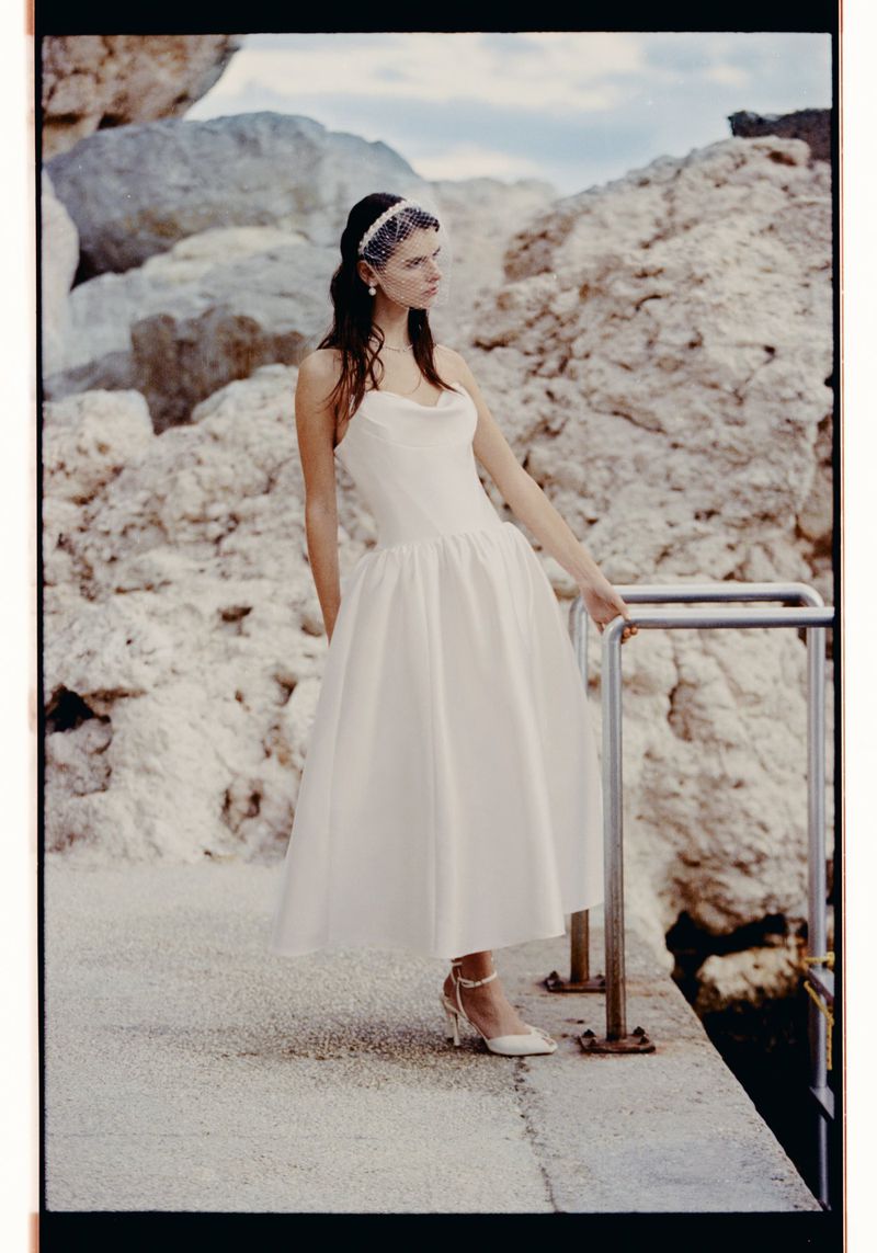 Woman in white gown stands by railing with rocky cliffs in the background, wearing a pearl headpiece and heeled shoes.