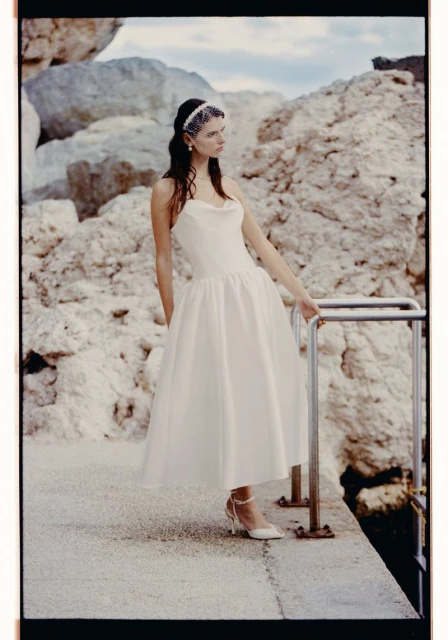 Woman in white gown stands by railing with rocky cliffs in the background, wearing a pearl headpiece and heeled shoes.