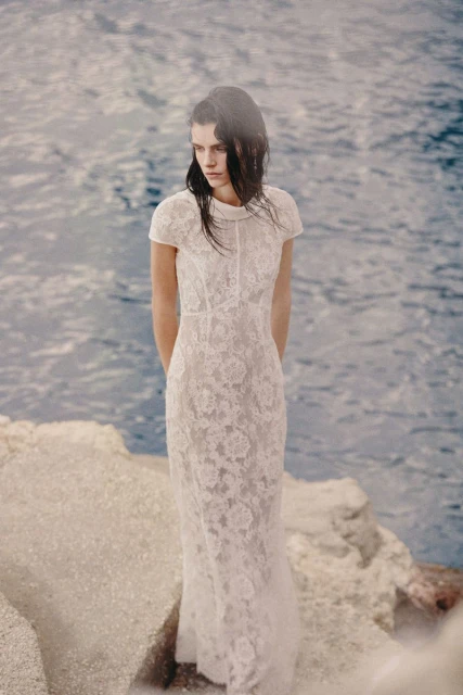 A woman in a white lace gown stands on rocks near the ocean with a serene expression, embodying fashion and bridal elegance.