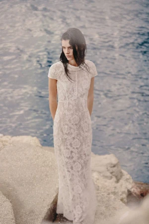 A woman in a white lace gown stands on rocks near the ocean with a serene expression, embodying fashion and bridal elegance.