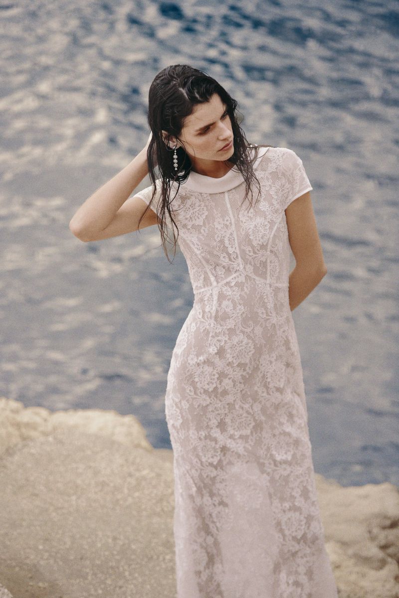 A slender woman with wet hair poses by the water's edge, wearing a long, white, lacy dress with a collared neckline, with the ocean behind her.