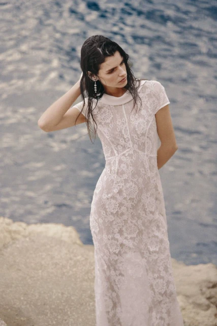 A slender woman with wet hair poses by the water's edge, wearing a long, white, lacy dress with a collared neckline, with the ocean behind her.