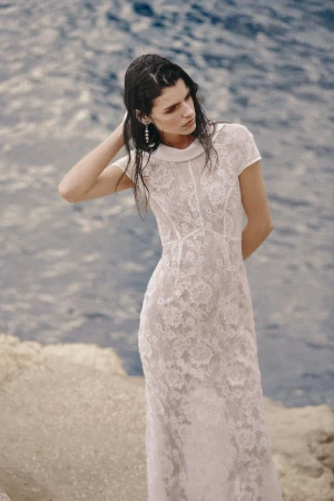 A slender woman with wet hair poses by the water's edge, wearing a long, white, lacy dress with a collared neckline, with the ocean behind her.