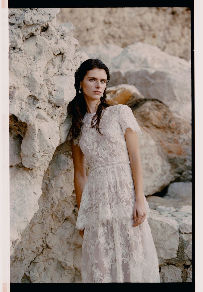 A woman poses in a lace dress against a rocky background. The light is soft, and the overall mood evokes natural beauty and elegance.