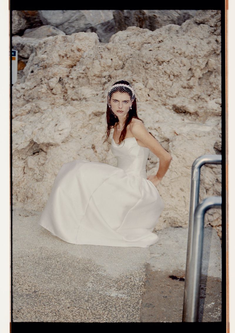 Woman in white wedding dress sits near a rock wall with water nearby. She wears a veil and has her hand on her hip.