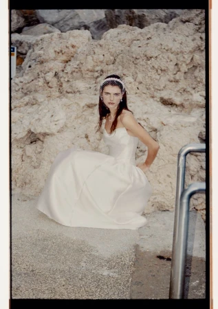 Woman in white wedding dress sits near a rock wall with water nearby. She wears a veil and has her hand on her hip.