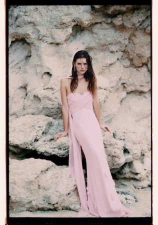 A young adult woman poses in a pale pink gown against a textured rock formation, styled for an outdoor fashion or beauty shoot.
