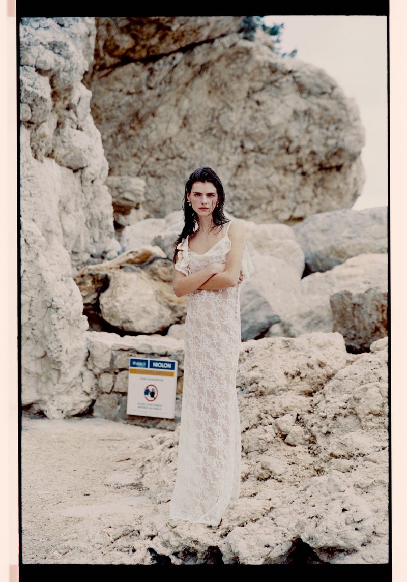 Woman in a white lace dress stands on a sandy beach with rock formations in the background, posing with crossed arms in outdoor setting.