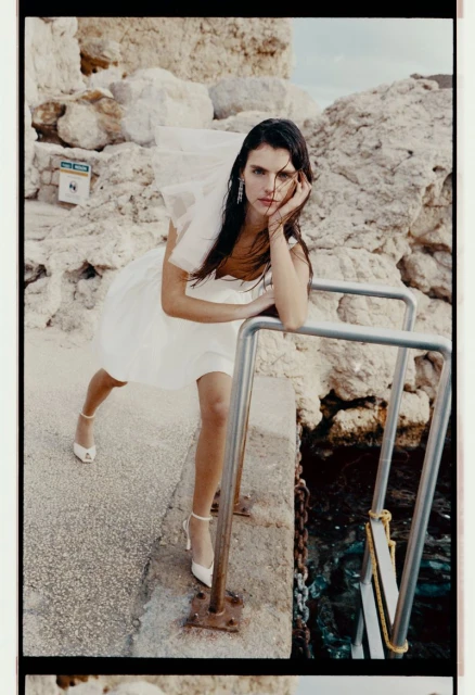 A woman in a white dress poses on a concrete structure by the ocean, with rocks visible in the background, captured in natural light.