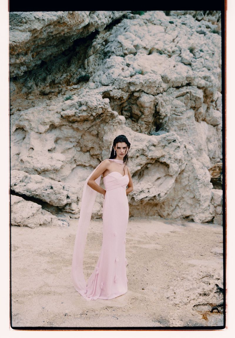 Woman in a pink dress poses against a rocky cliff backdrop. The photograph captures a blend of fashion and natural landscape.