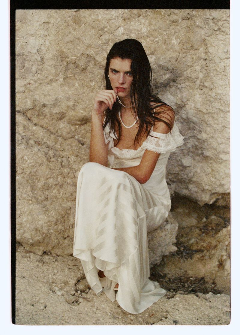 Adult woman with wet hair sitting in a long white dress against a light rock face. She wears a pearl necklace and a neutral facial expression.