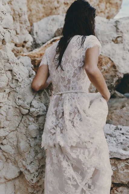 Woman in white lace dress stands on a rocky beach, back turned, wet hair, facing stone cliffs in soft daylight. Fashion shot.