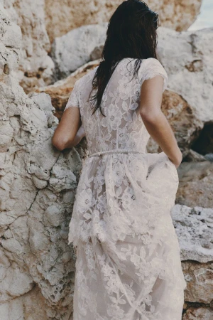Woman in white lace dress stands on a rocky beach, back turned, wet hair, facing stone cliffs in soft daylight. Fashion shot.