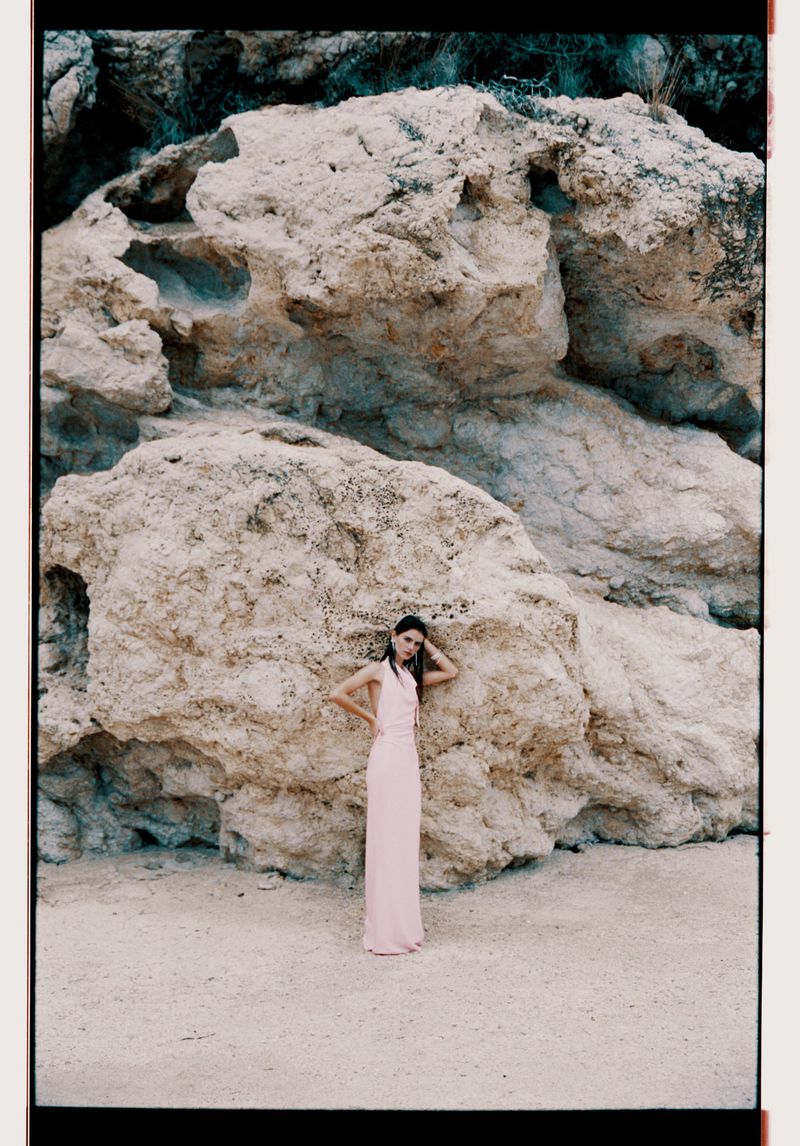 A woman in a light pink dress poses in front of a large rock formation on a sandy surface, fashion photography, outdoor setting.