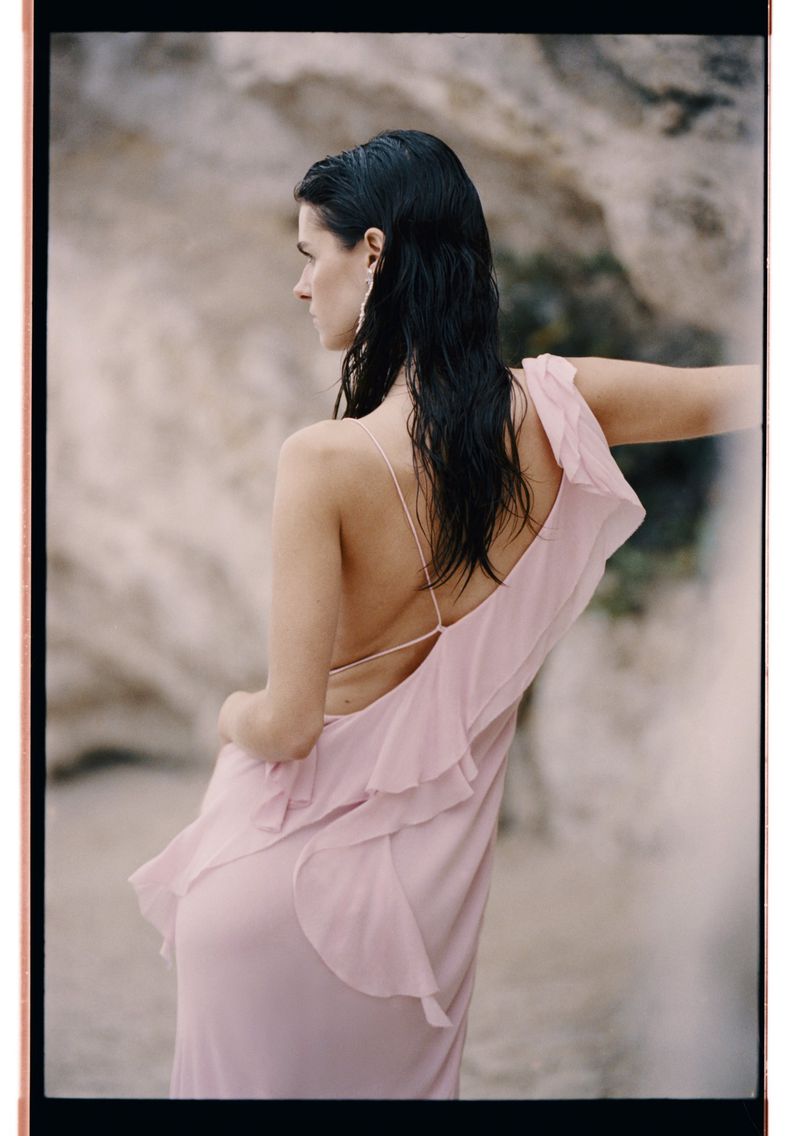 Woman in flowing pink dress poses against a rocky backdrop, showcasing fashion and elegance in a natural setting. Wet hair, bare back.