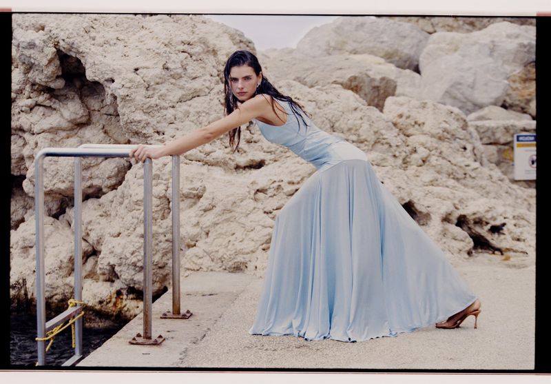 Adult female fashion model in a long blue dress posing on a concrete pier with a rock and metal railing seaside backdrop.