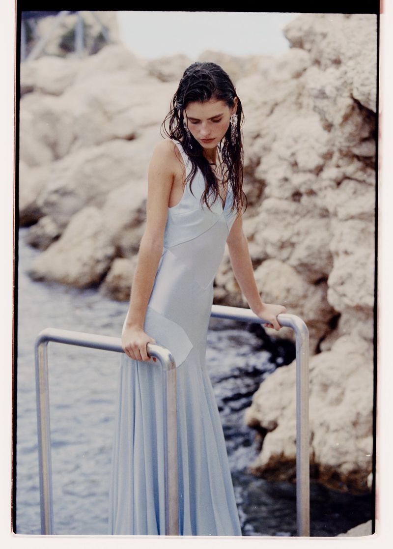 A woman with wet hair, wearing a light blue satin dress, poses near a rocky beach and calm water, with a metallic railing.