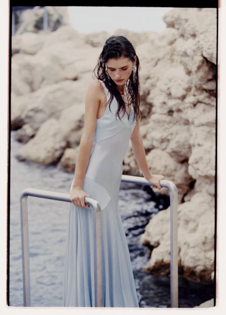 A woman with wet hair, wearing a light blue satin dress, poses near a rocky beach and calm water, with a metallic railing.