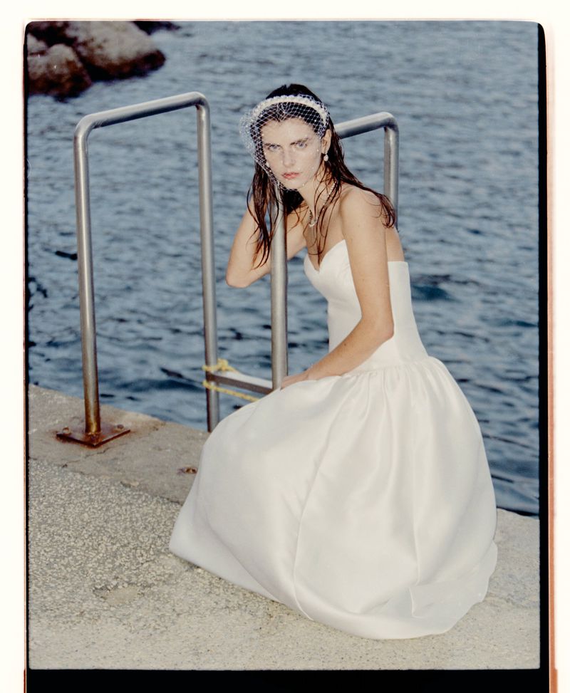 Woman in white strapless gown and bridal veil sits on concrete near metal railing with the ocean in the background on a sunny day.