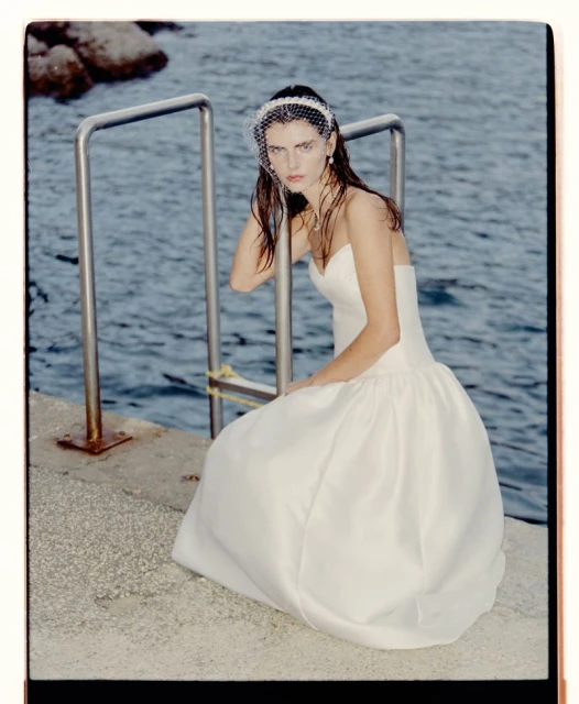 Woman in white strapless gown and bridal veil sits on concrete near metal railing with the ocean in the background on a sunny day.