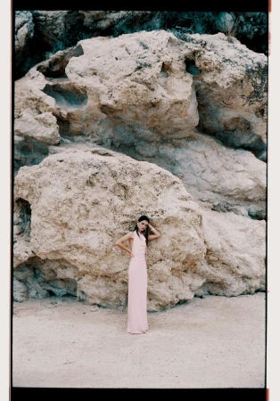 Model standing in a long pink dress against a large rock formation and sandy ground