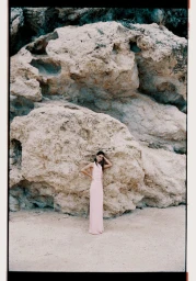 Model standing in a long pink dress against a large rock formation and sandy ground