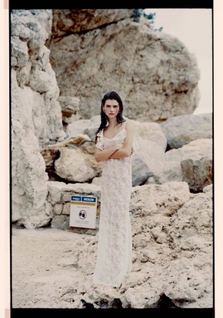 Model in a white lace dress standing outdoors against a rocky coastal cliff formation