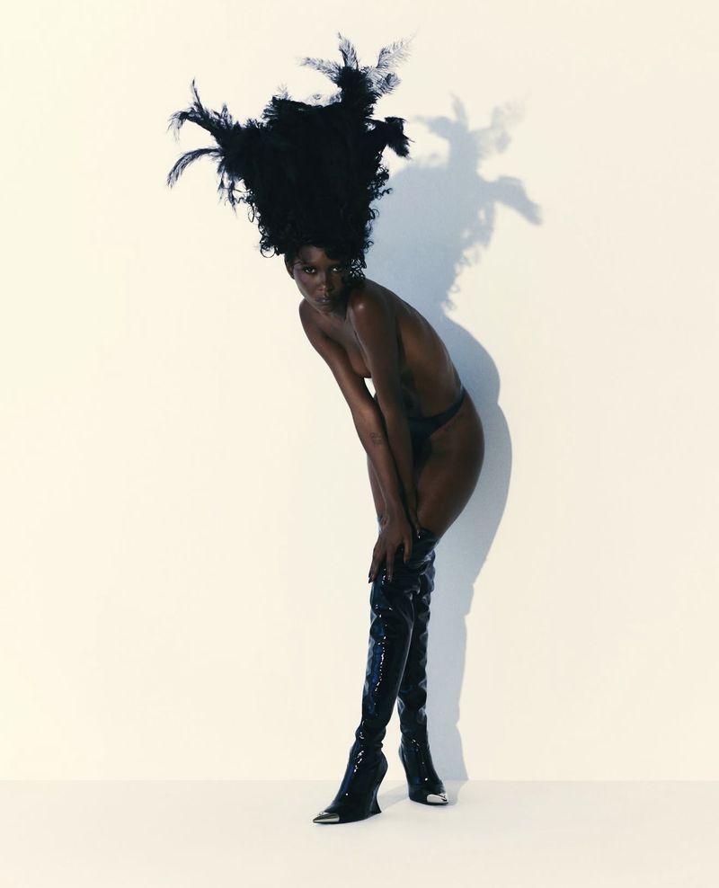 Striking fashion portrait of a bare-chested black woman in thigh-high patent boots and large feather headdress, standing against a white backdrop.