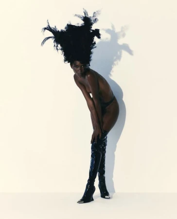 Striking fashion portrait of a bare-chested black woman in thigh-high patent boots and large feather headdress, standing against a white backdrop.