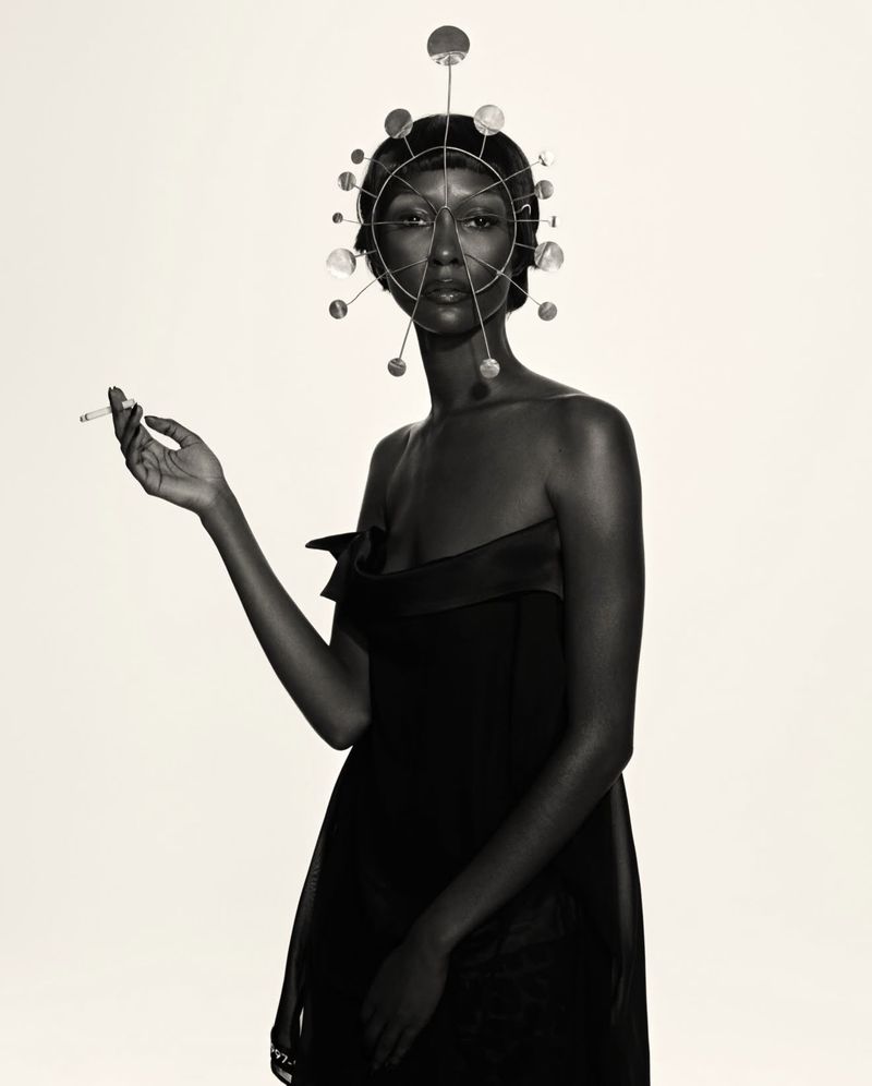 Black and white portrait of a woman in a black dress holding a cigarette, adorned with a circular, silver headpiece.