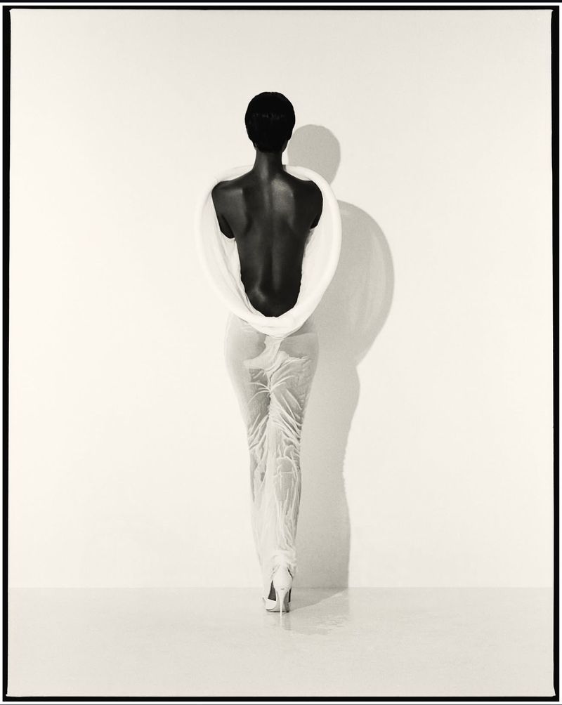 Elegant, back-facing view of a slender African-American woman in light clothing and high heels, standing against a bright background.