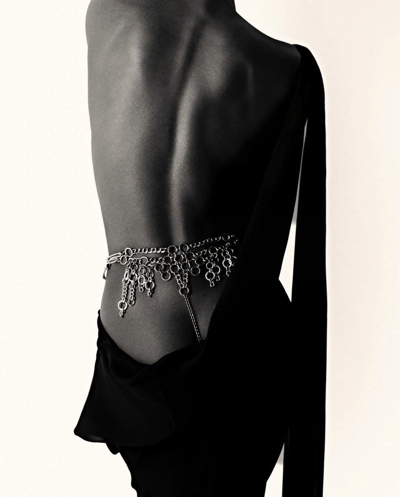 Back view of a slender adult female with a silver chain belt, draped over a black dress against a plain off-white background in a studio.