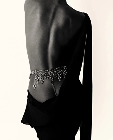 Back view of a slender adult female with a silver chain belt, draped over a black dress against a plain off-white background in a studio.