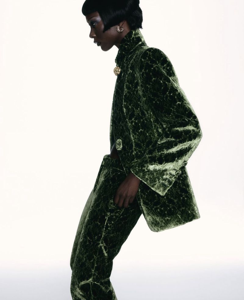 An adult female model with short dark hair poses in a green, patterned velvet blazer and pants against a white background.