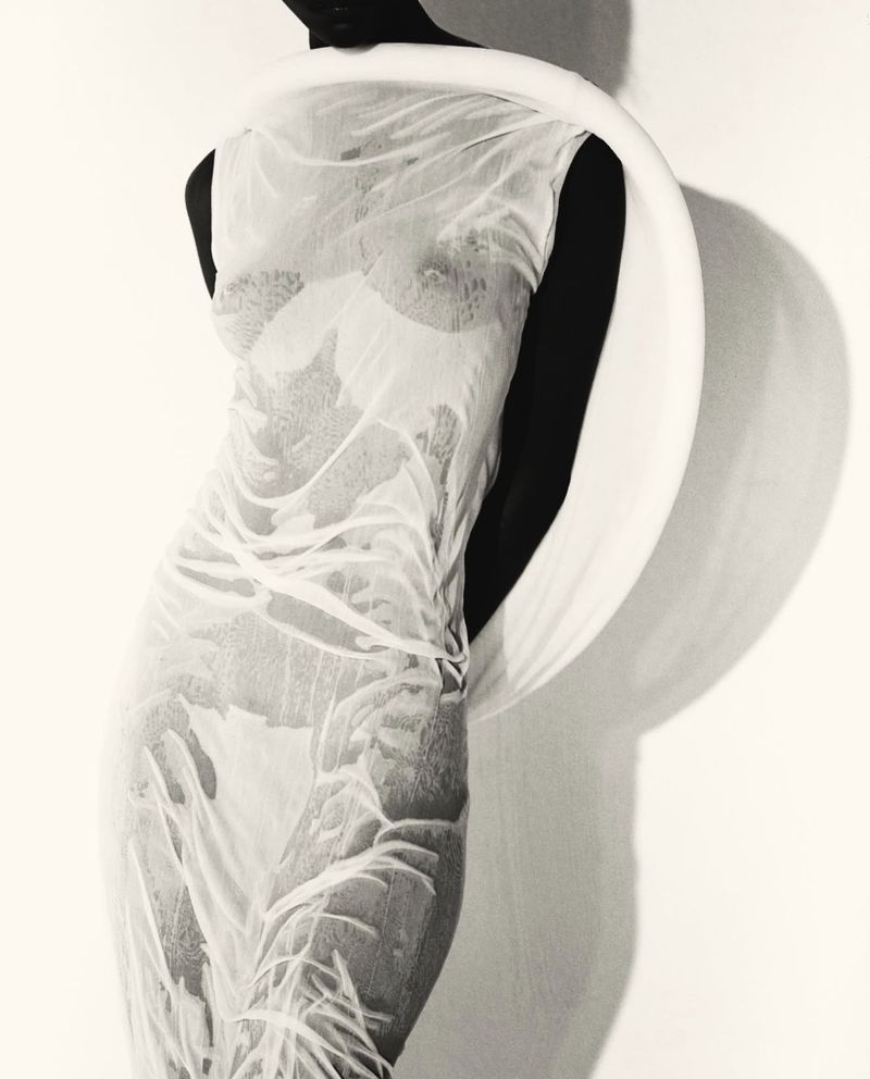 Monochromatic fashion shot of a dark-skinned woman in a flowing white dress, standing against a white wall with shadows, artistic lighting.